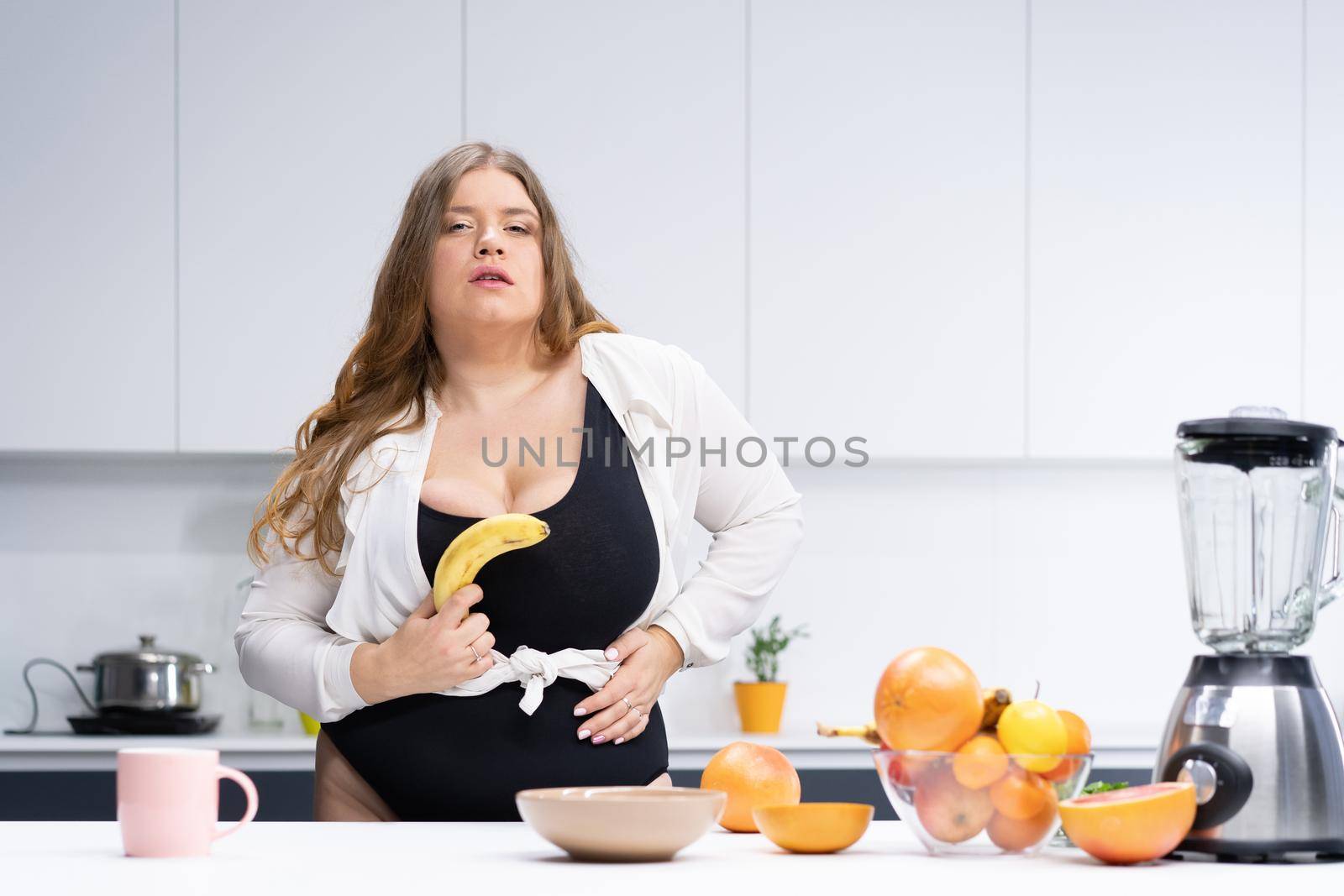 Dieting and nutrition concept. Happy curvy body young woman with long blond hair using blender at modern kitchen, blending fresh fruits for healthy smoothie.