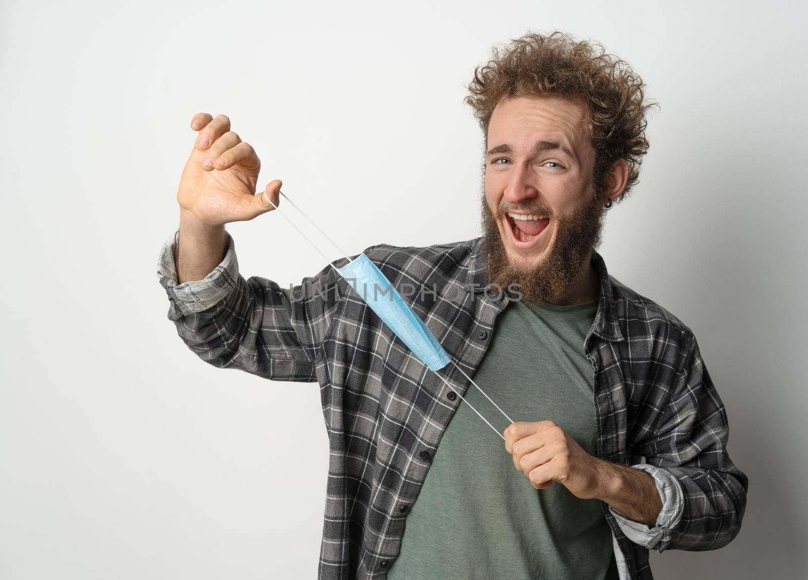 Fooling around playing with protective medical face mask young handsome man holding it stretching it in front of face wearing plaid shirt and olive t-shirt under. White background by LipikStockMedia