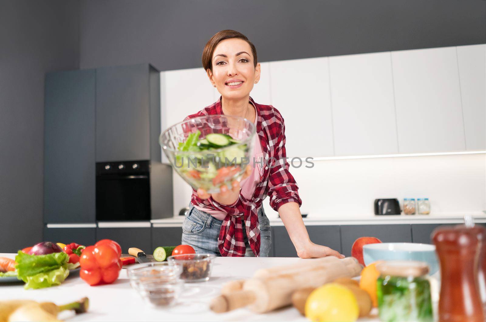 Happy young housewife holding bawl of salad showing it on camera prepared for a family dinner or girls night standing in the kitchen. Healthy food living. Healthy lifestyle.