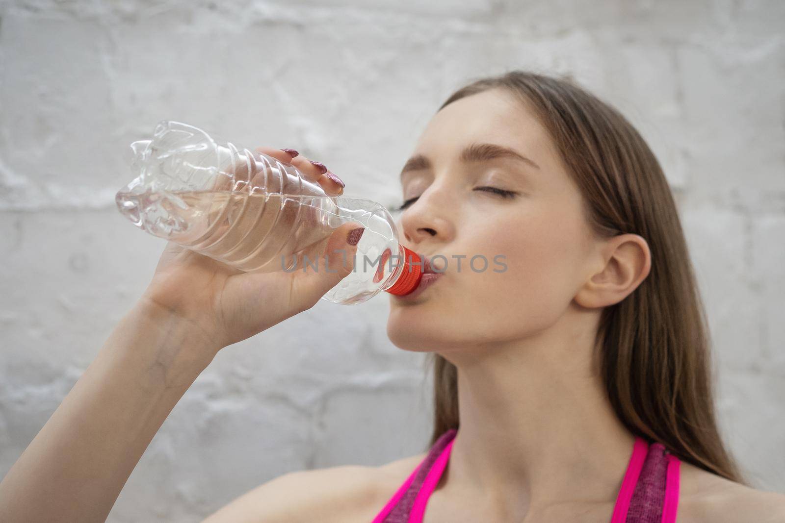 Young athlete woman drink water from plastic bottle while takes a break. Beautiful athletic girl In pink color sportswear fitness workout on white wall Background. High Quality Image.