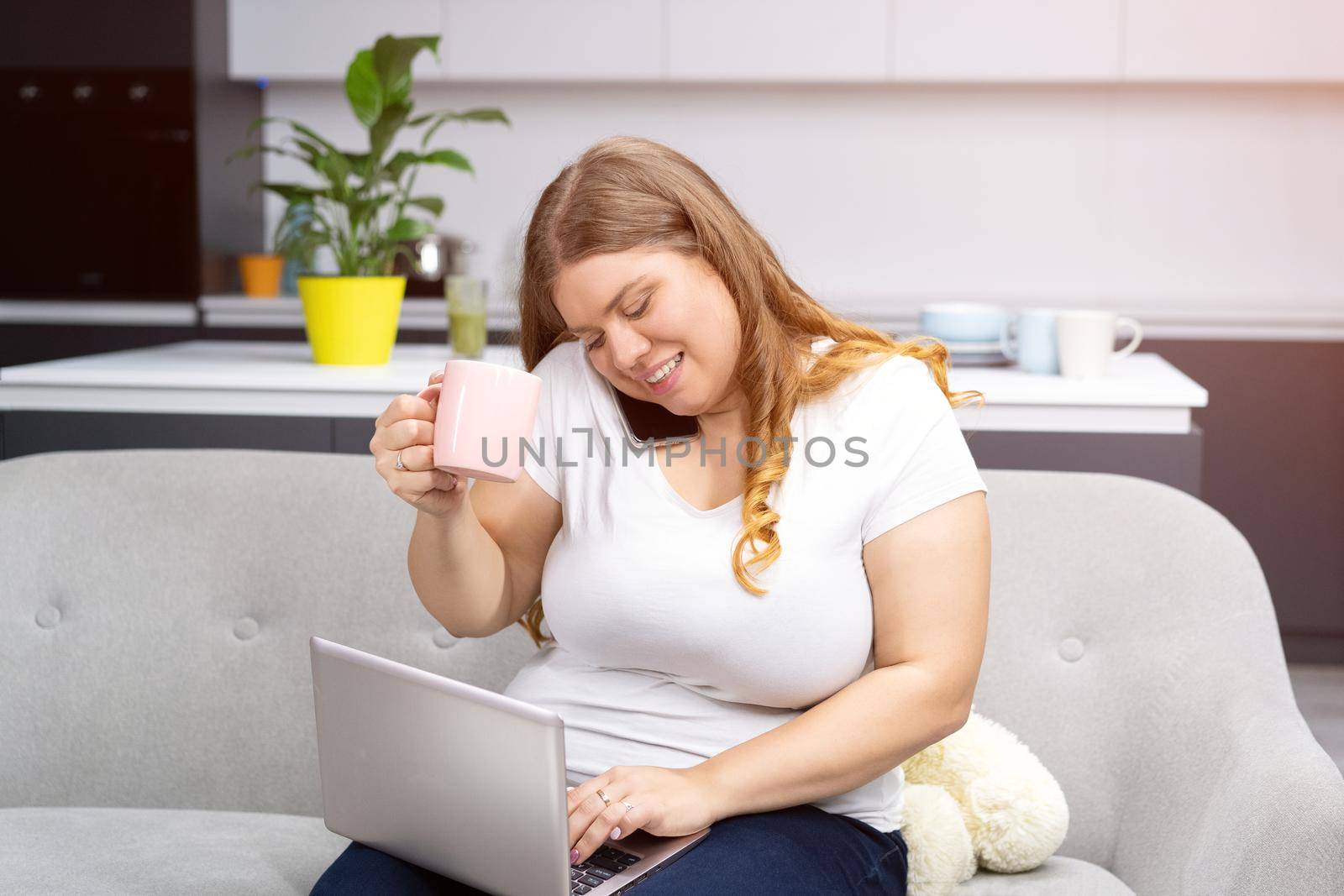 Drinking coffee or tea while working on laptop sitting at home curve body young woman staying at home during quarantine. Self isolation as prevention. Working distantly from home using laptop.