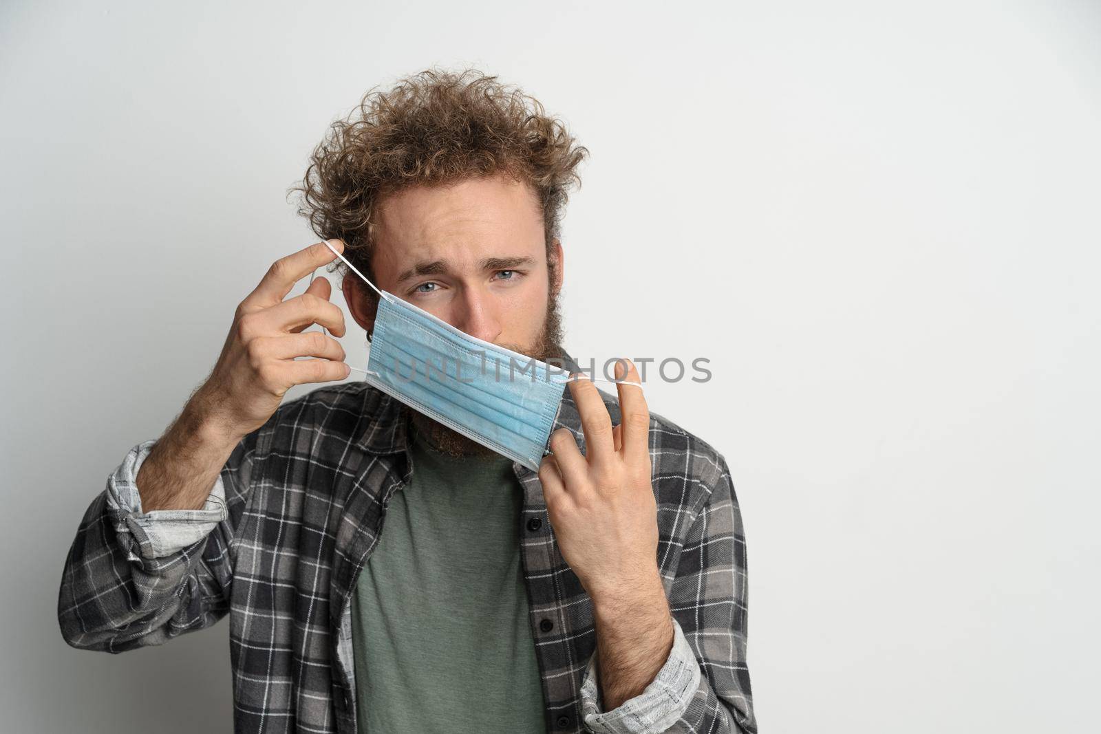 Young man holding protective sterile medical mask in front of face to protect coronavirus, with curly hair, wearing plaid shirt and olive t-shirt under. White background.