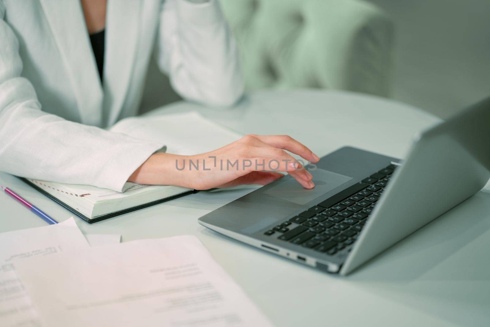 No face visible Businesswoman working sitting in front of laptop wearing white official suit. Office worker female using touchpad browsing laptop while sitting at her working place. Close up by LipikStockMedia