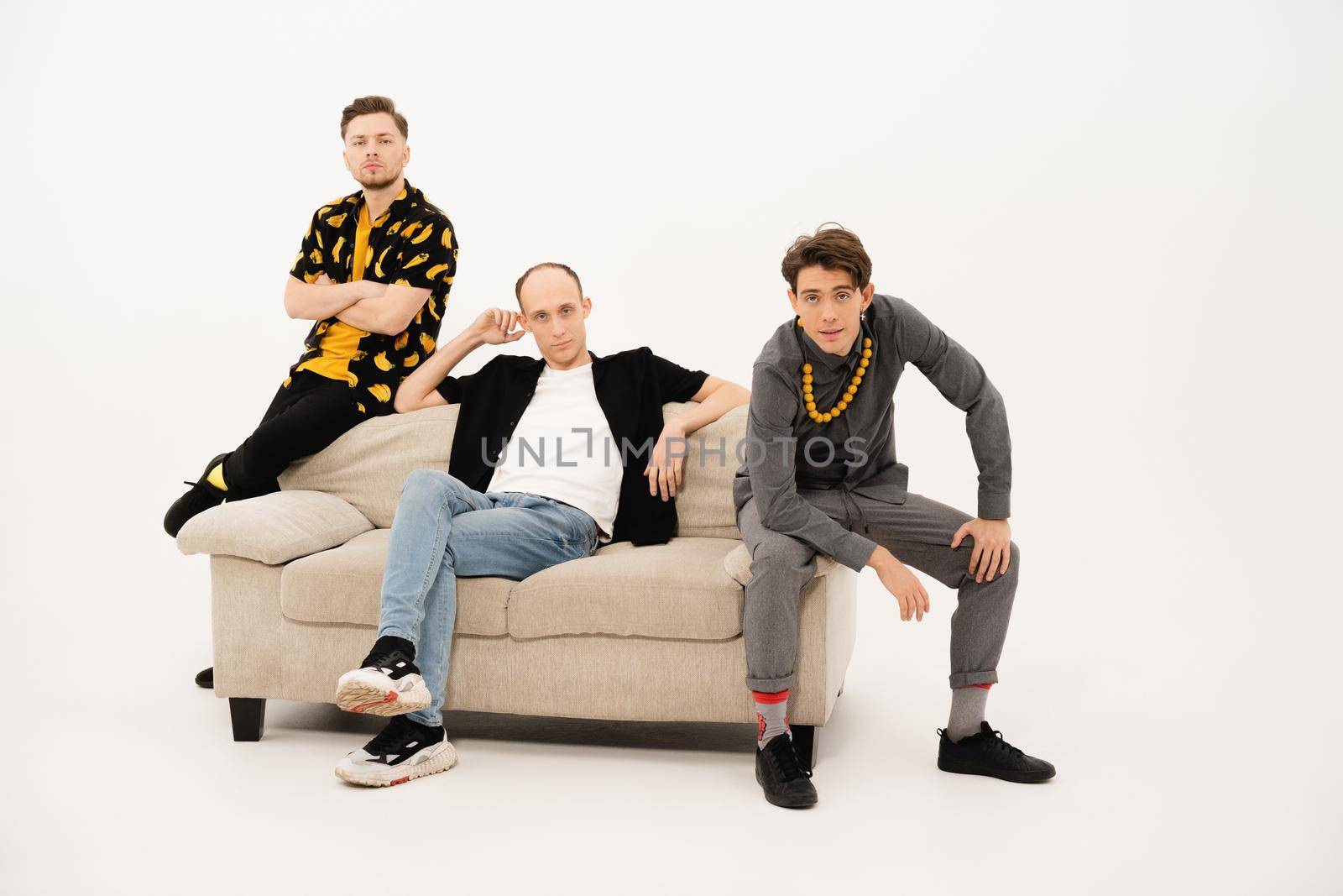 Friends seated on couch in studio. Three stylish young guys talking on the camera. Group of friends are sitting on a soft couch and communicates isolated on white background by LipikStockMedia