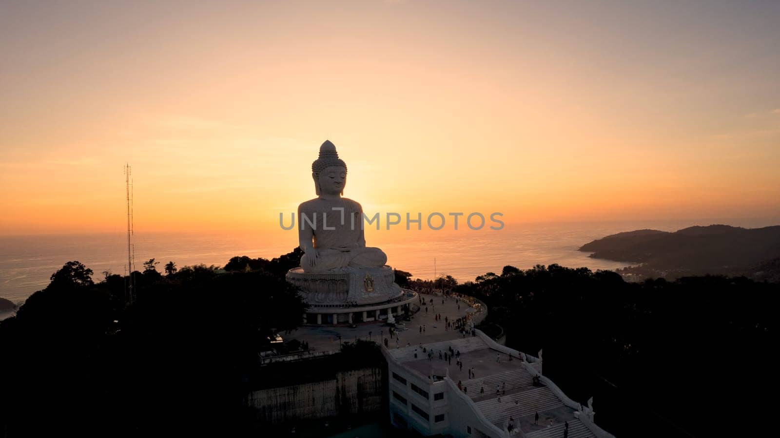 Big Buddha at sunset view from a drone. Phuket. People climb the steps to the statue. The green hills of the island are all around. In the distance, the bright sun goes over the sea. View from above