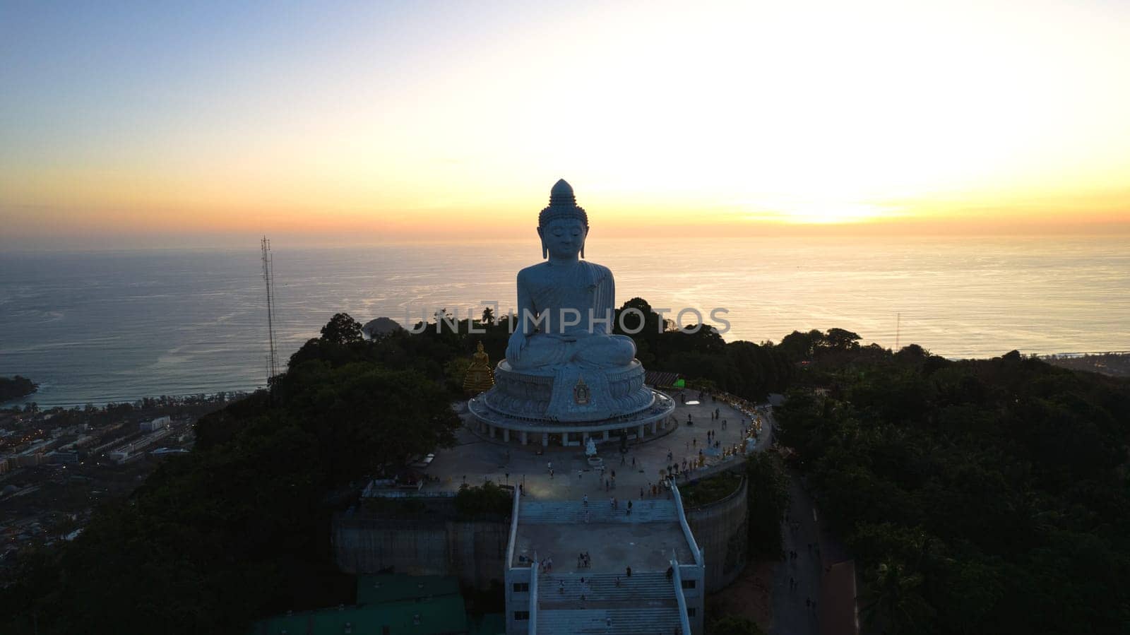 Big Buddha at sunset view from a drone. Phuket. People climb the steps to the statue. The green hills of the island are all around. In the distance, the bright sun goes over the sea. View from above