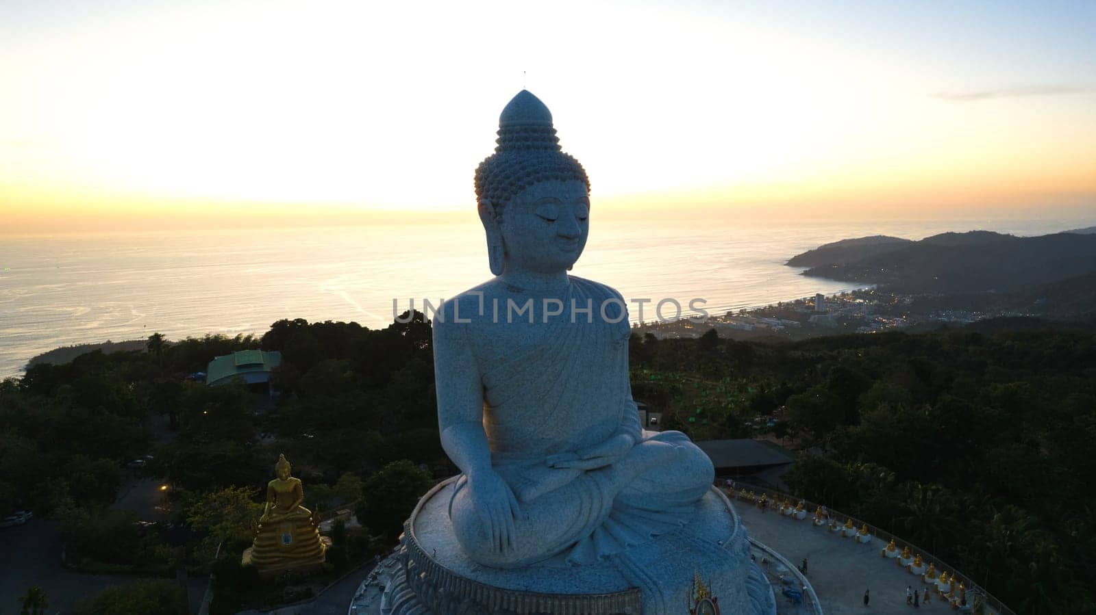 Big Buddha at sunset view from a drone. Phuket. People climb the steps to the statue. The green hills of the island are all around. In the distance, the bright sun goes over the sea. View from above