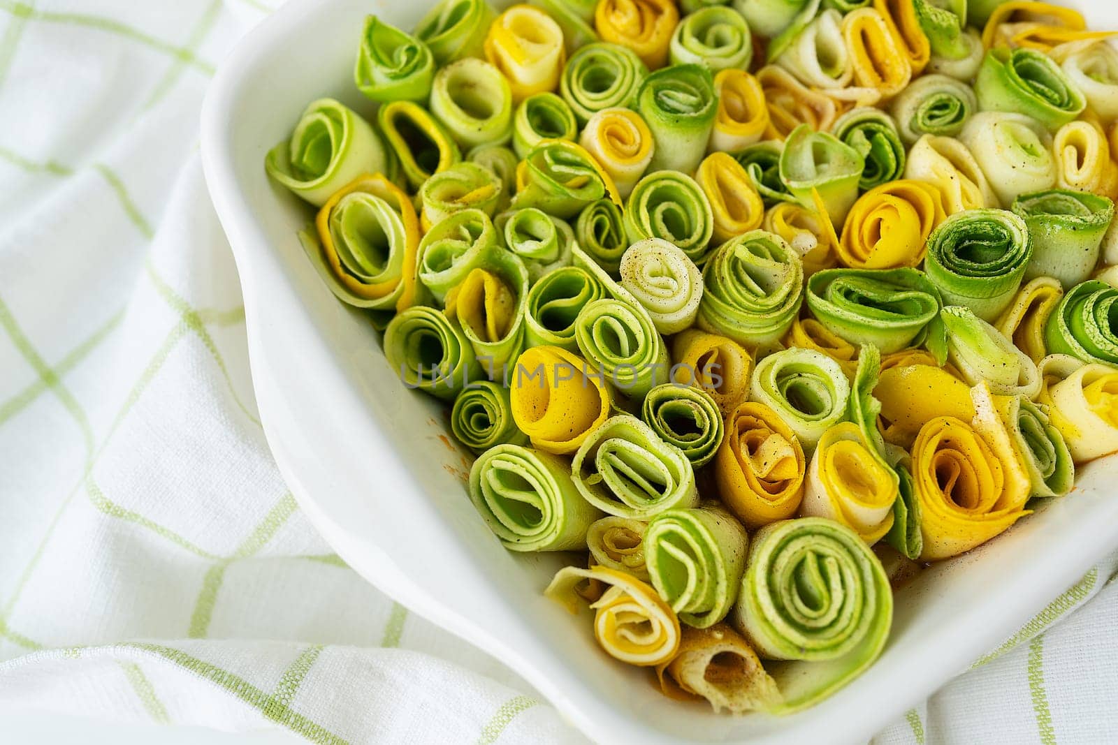 Small rolls of zucchini green and yellow with creamy sauce in a white baking dish against the background of a checkered napkin