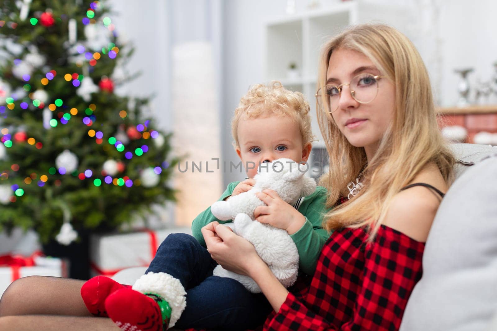 The woman is wearing an elegant plaid dress. The woman is holding a small child on her lap. Both are sitting on a gray couch. The room is decorated with Christmas decorations. The woman is wearing corrective glasses.