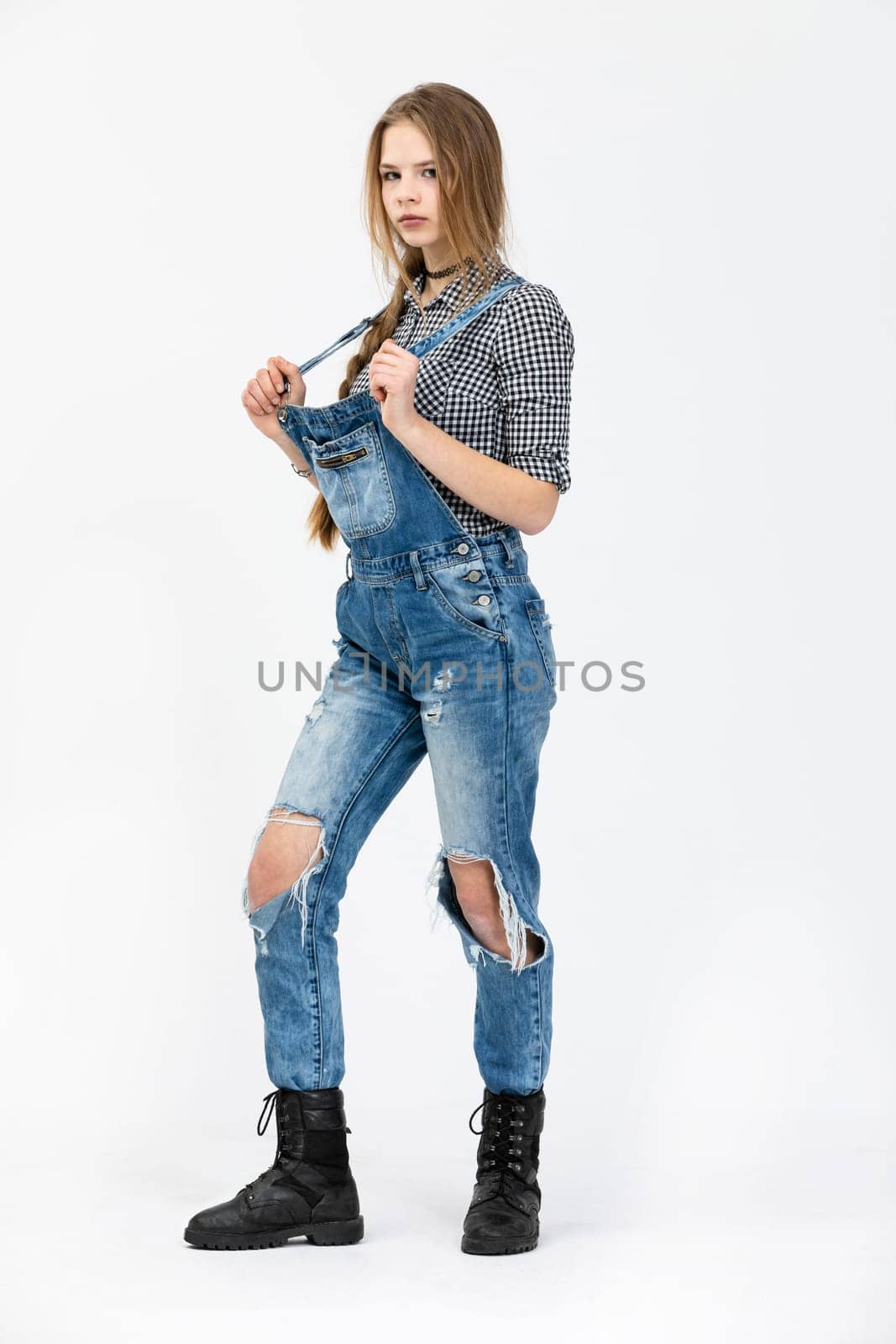 A young girl poses for a photo in a standing position., The girl is wearing a checked shirt and rubbed jeans. The girl is wearing black high top shoes. She is styled in a braid and has disheveled hair.