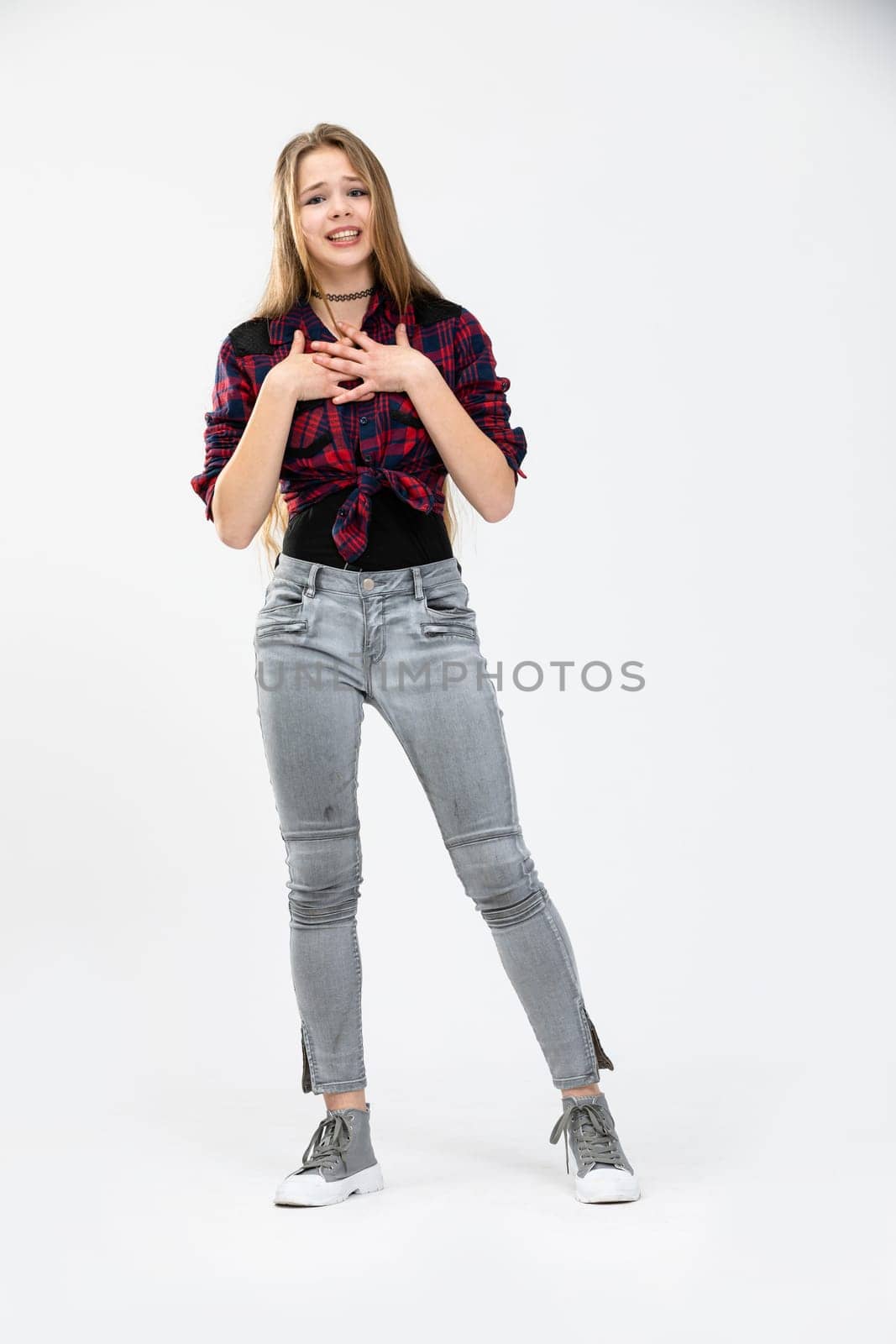 A long-haired girl with loose hair stands against a white background. The woman presses her hands to her chest. The girl has a grimace on her face and looks unhappy.