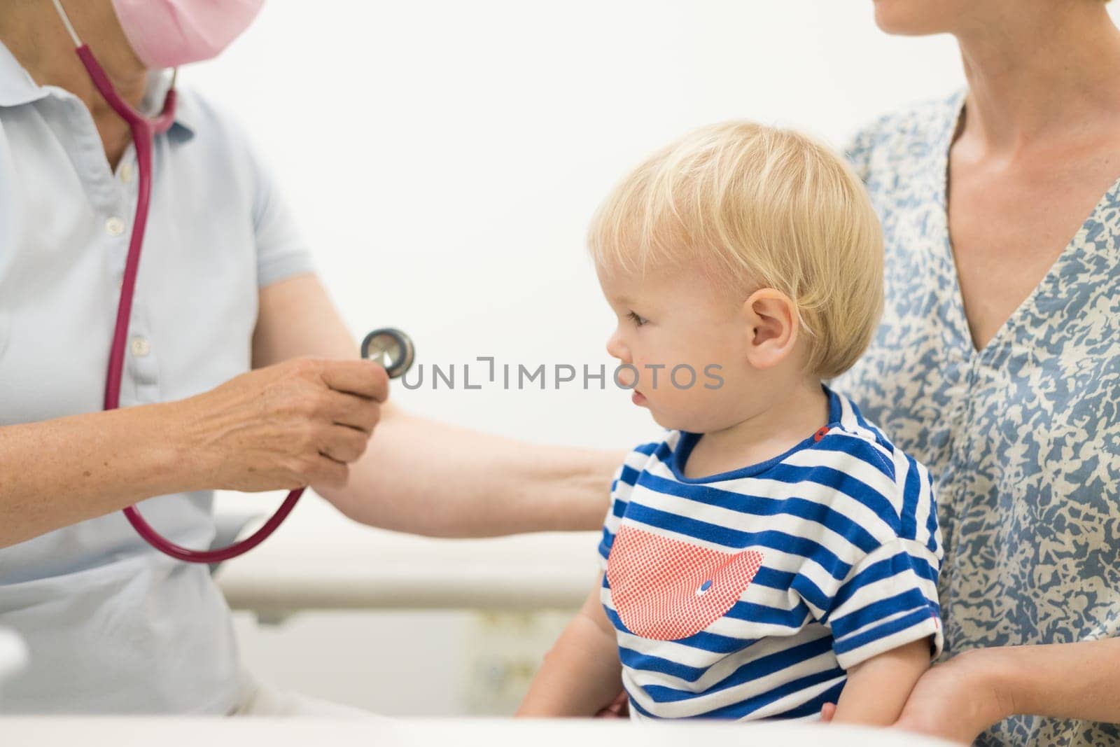 Infant baby boy child being examined by his pediatrician doctor during a standard medical checkup in presence and comfort of his mother. National public health and childs care care koncept