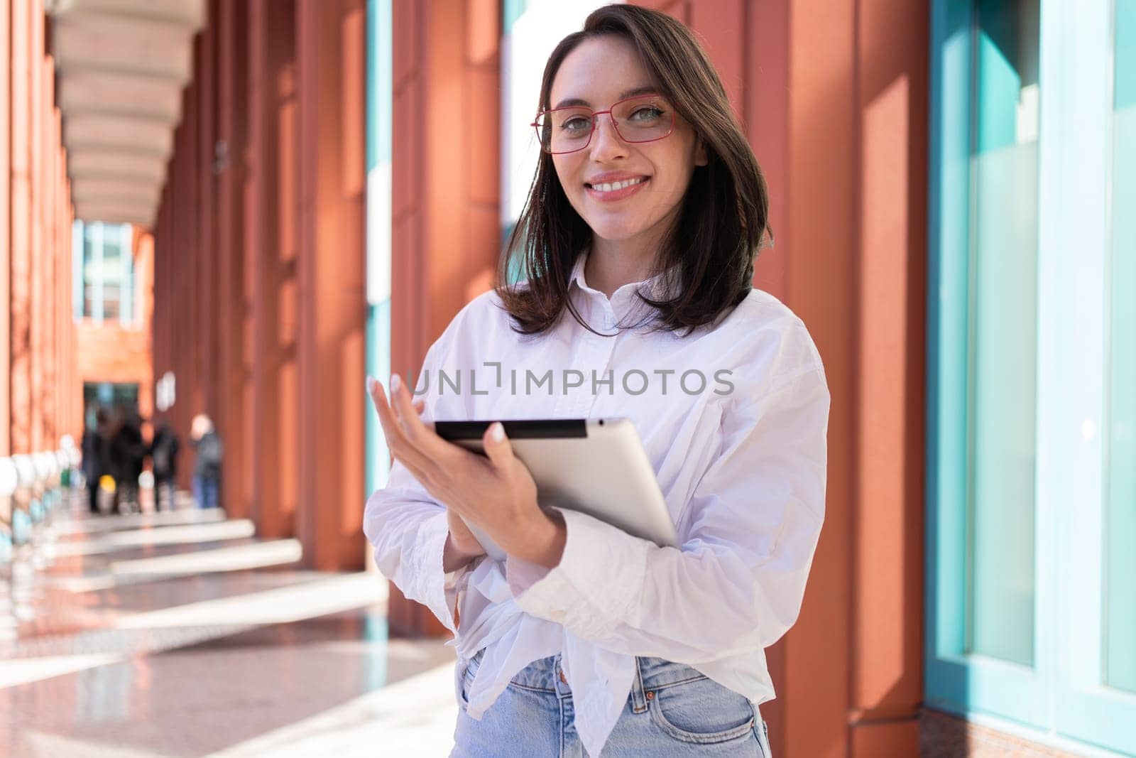 Businesswoman in glasses using digital tablet touching screen looking at camera, standing outdoor dressed white shirt. Female freelancer holding tablet while walk outside