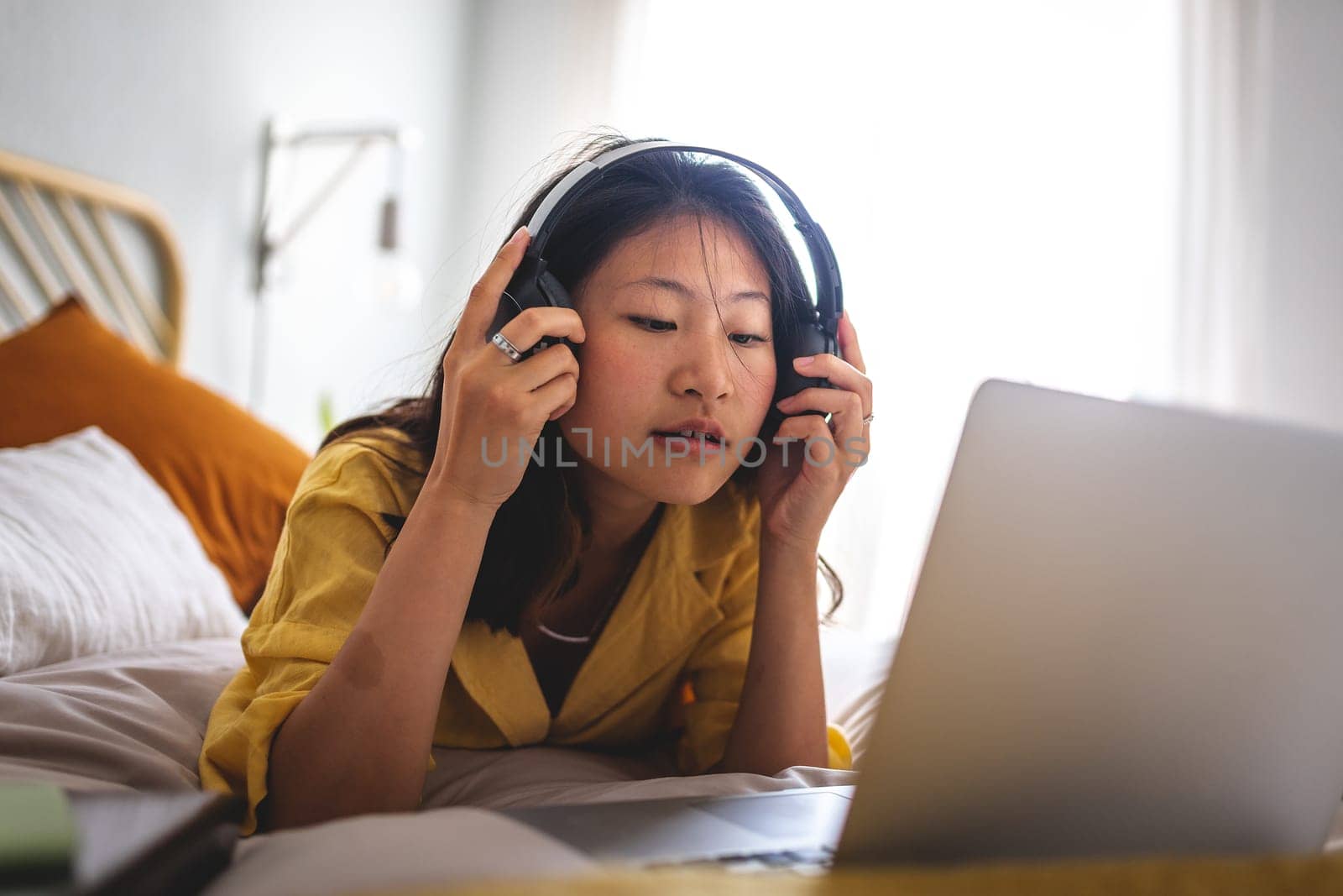 Teen asian girl putting on headphones to attend online classes. Chinese female watch movie on laptop. Listen to music at home lying on bed. Lifestyle and e-learning concept.