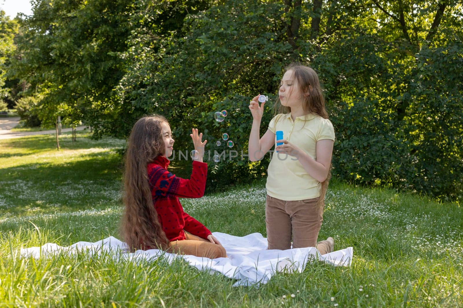 Joyful Asian Caucasian Girls, Children Of Elementary School Age Blow Soap Bubbles In Summer Park, Meadow. Happy Childhood. School Break Time. Children Protection Day, Earth Day. Horizontal Plane by netatsi