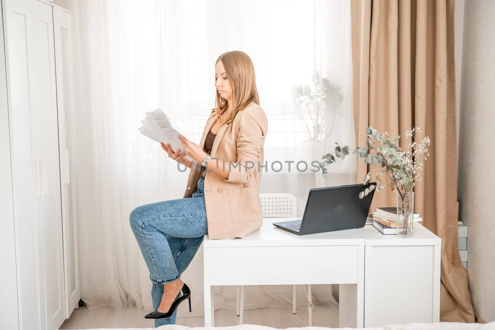 Business woman sitting on desk in home office, positive female lawyer rejoices throwing papers up. She is wearing a beige jacket and jeans