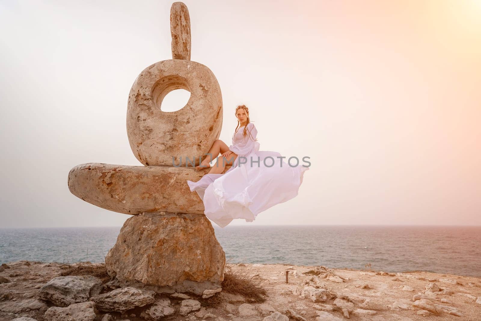 Woman sea stone. A woman stands on a stone sculpture made of large stones. She is dressed in a white long dress, against the backdrop of the sea and sky. The dress develops in the wind