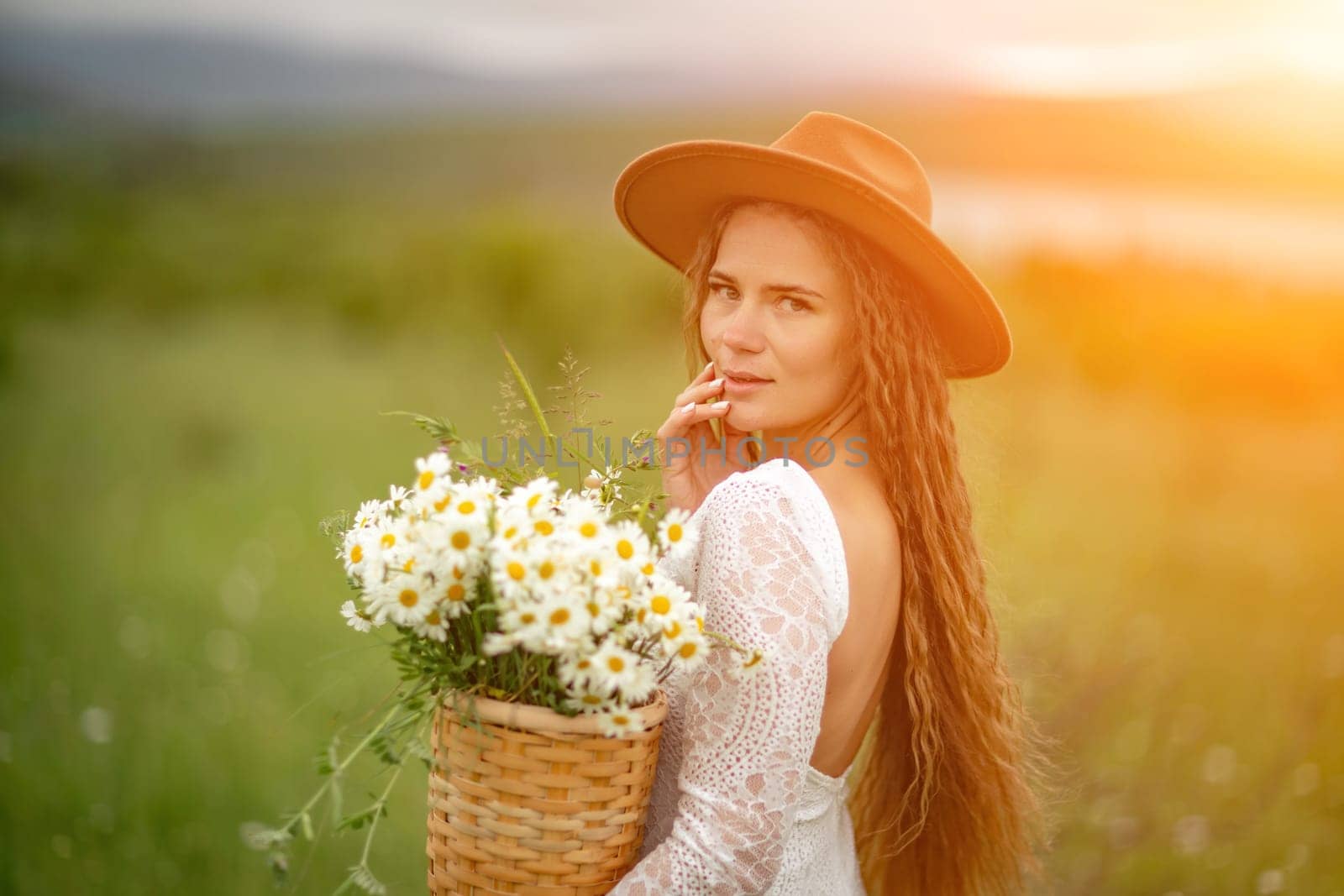 A middle-aged woman in a white dress and brown hat stands on a green field and holds a basket in her hands with a large bouquet of daisies. In the background there are mountains and a lake