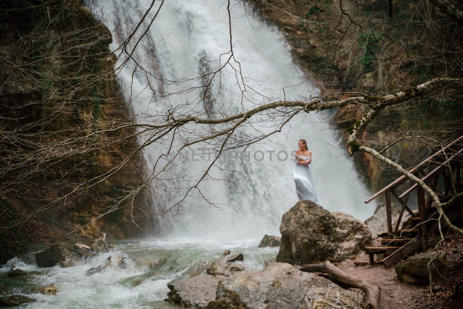 Happy woman in a white dress stands on a stone with a waterfall behind.