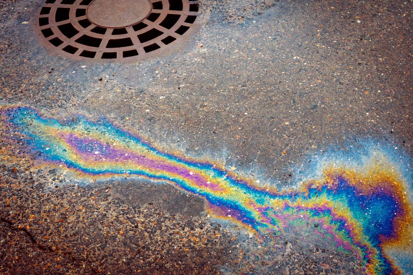 Oil slick on the asphalt road background drains into the storm drain. Abstract background from motor oil, gas or petrol spilled on asphalt.