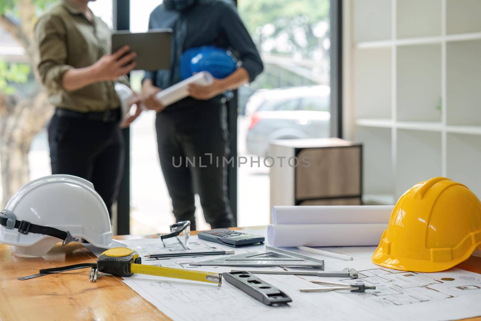Concept of engineering equipments of protective helmet for competent engineer on piles of paper works. Blurred image of engineer working together at the desk.