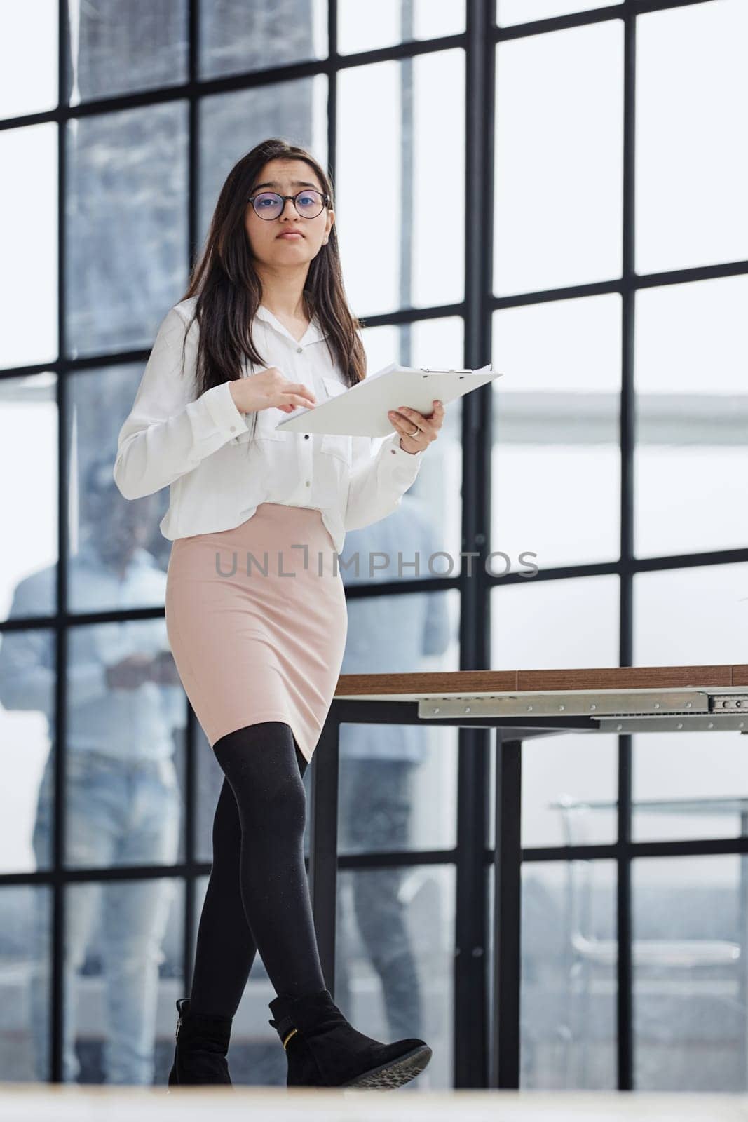 Beautiful confident young busines woman with black hair in a modern office standing near the table with a folder in her hand by Prosto