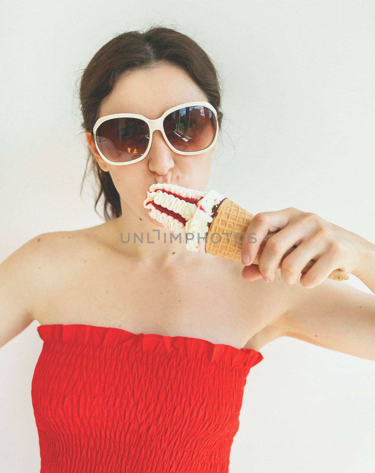 Cinematic photo filter. Happy cheerful girl in a red top and white sunglasses eats a colorful ice cream cone in her hands and smiles. by sfinks