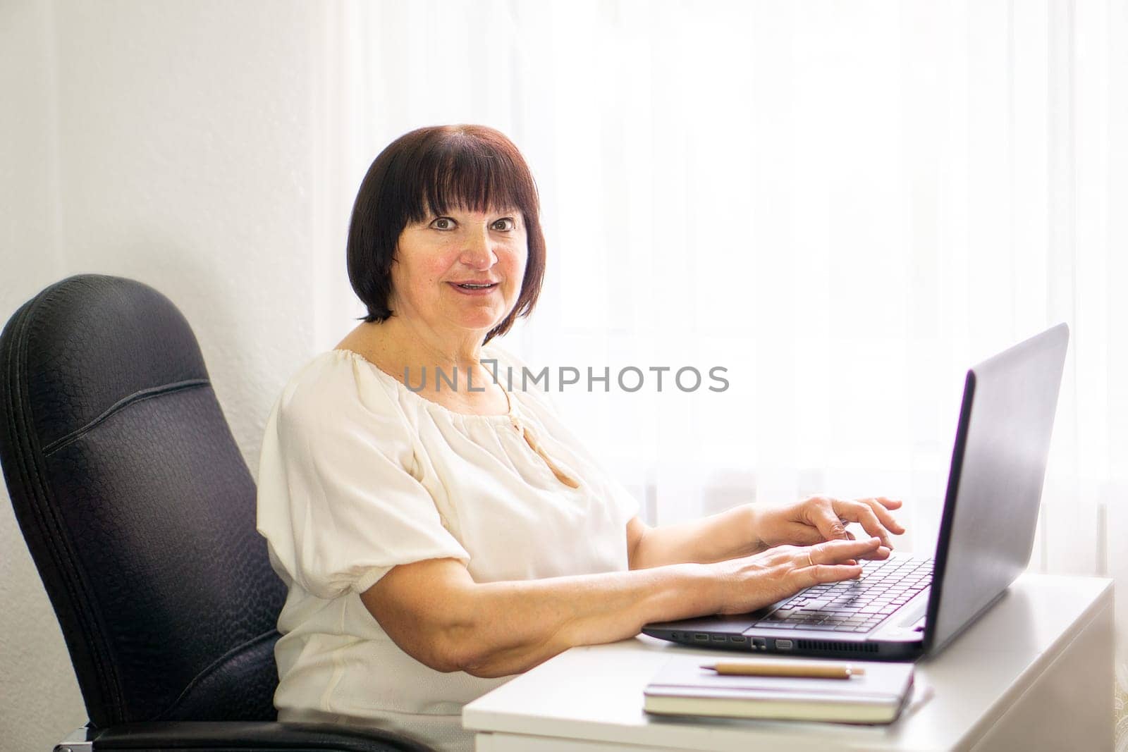 Senior smiling business woman working at her computer laptop at home. Business, education and technology concept. Portrait of a skilled female programmer freelancer with laptop sitting on the table in an office.