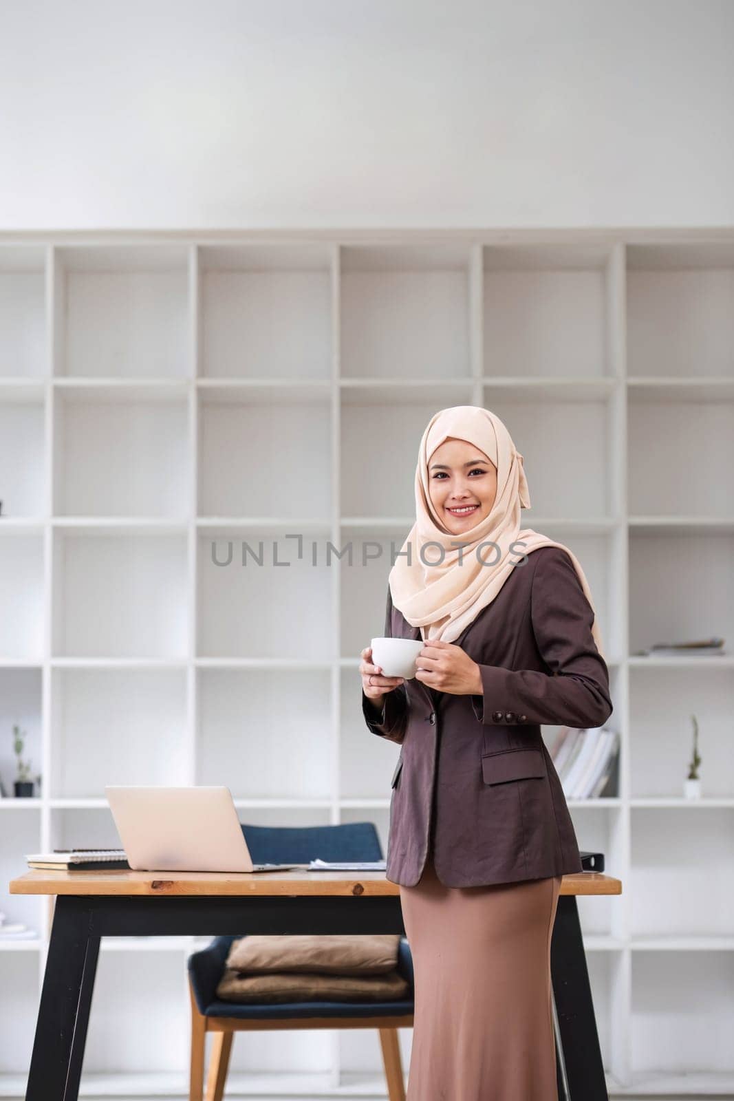 A portrait of a confident and smiling Asian Muslim businesswoman stands in her modern office with a coffee cup in her hands. by wichayada