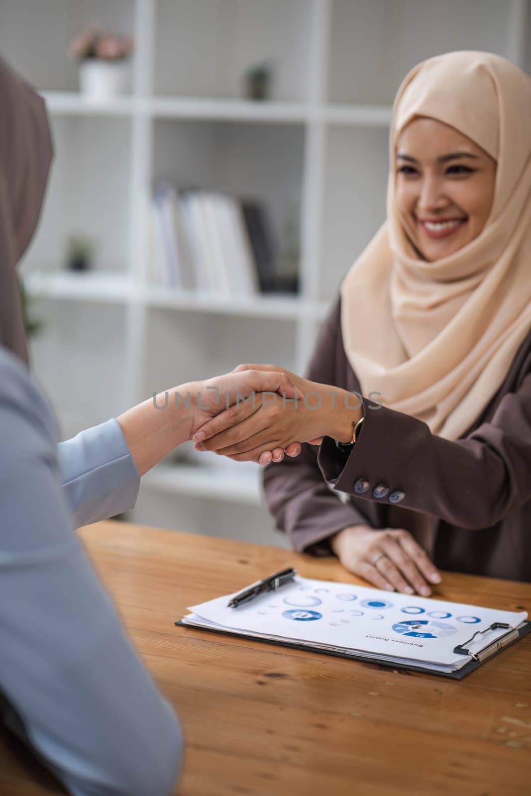 A beautiful and cheerful Asian Muslim businesswoman shaking hands with a female business partner during the meeting in the office. by wichayada