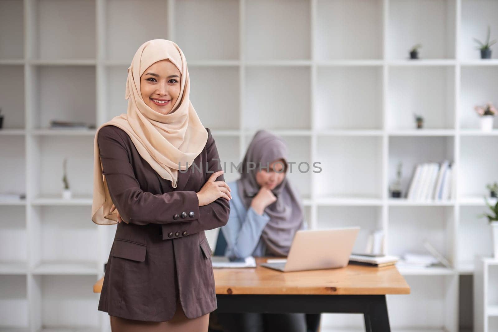 A portrait of a confident and smiling Asian Muslim businesswoman stands in her modern office with a coffee cup in her hands...