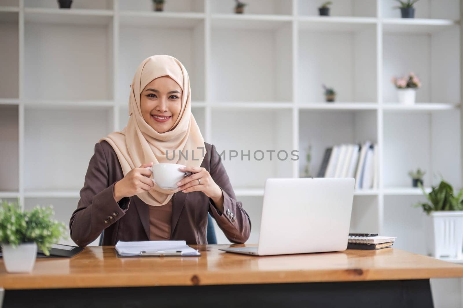 A portrait of a beautiful and successful Asian Muslim businesswoman or female boss in hijab, smiling and looking at the camera while sitting at her desk with a coffee cup in her hands. by wichayada