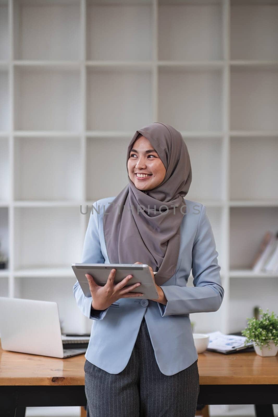 A portrait of a successful Asian Muslim businesswoman stands in her modern office with a digital tablet in her hand. by wichayada