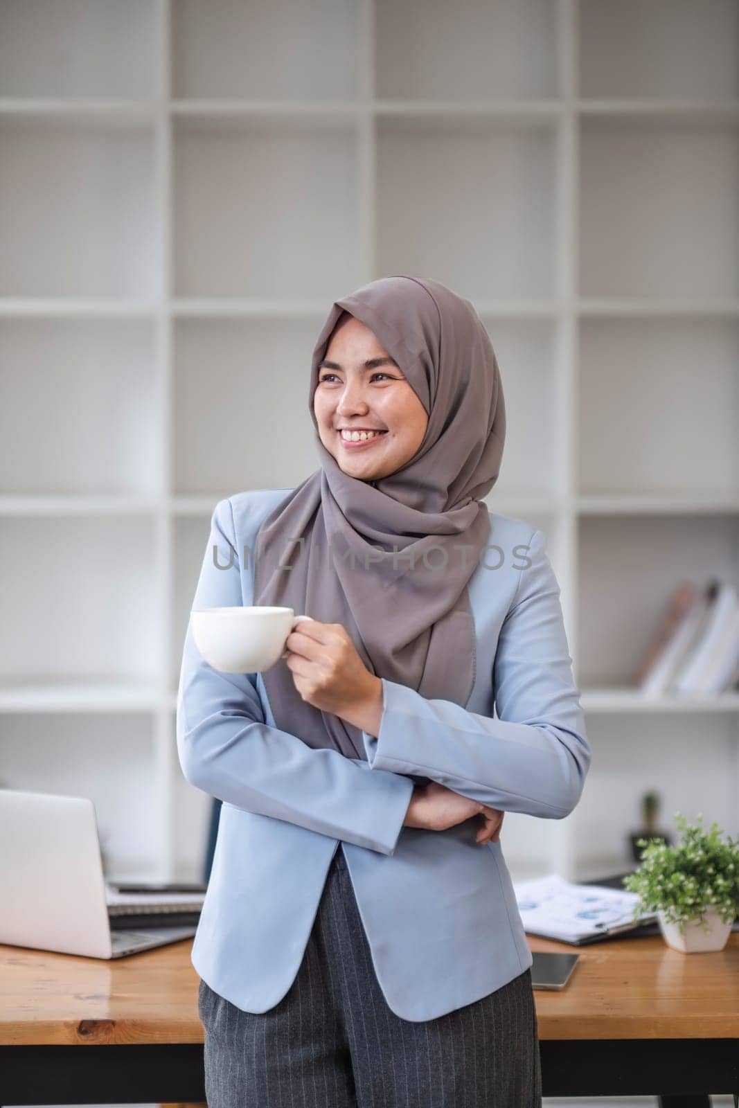 A portrait of a confident and smiling Asian Muslim businesswoman stands in her modern office with a coffee cup in her hands. by wichayada