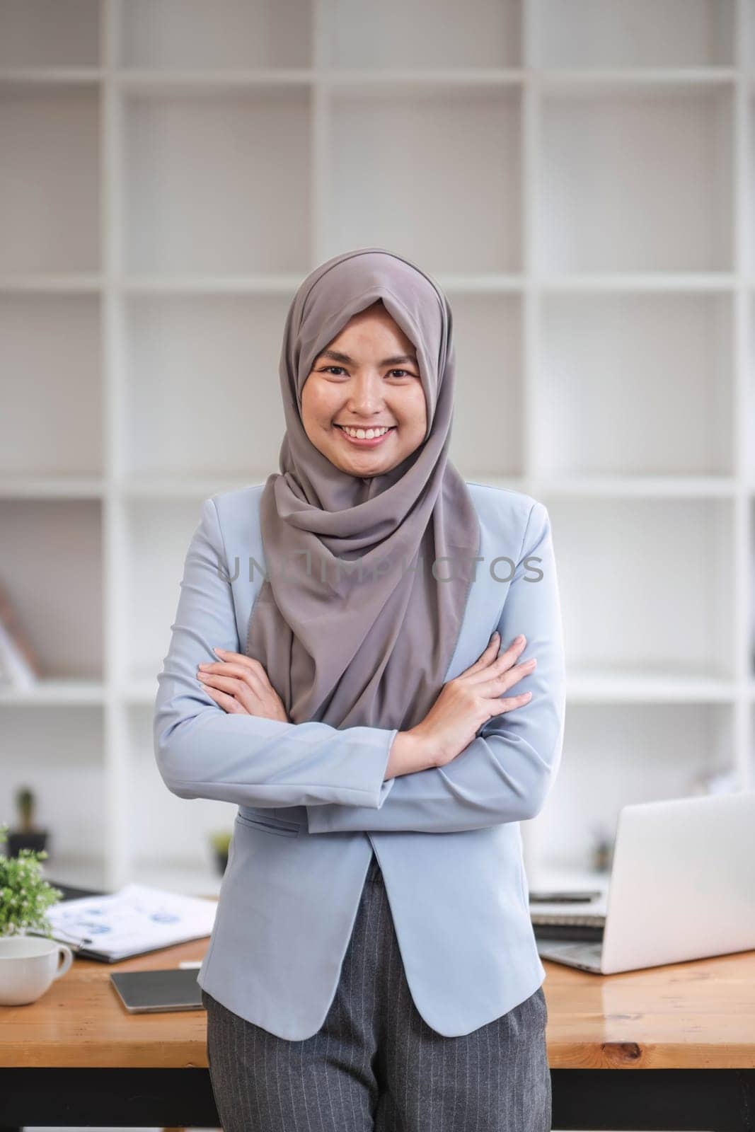 A portrait of a beautiful and professional Asian Muslim businesswoman stands in the office with her arms crossed. by wichayada