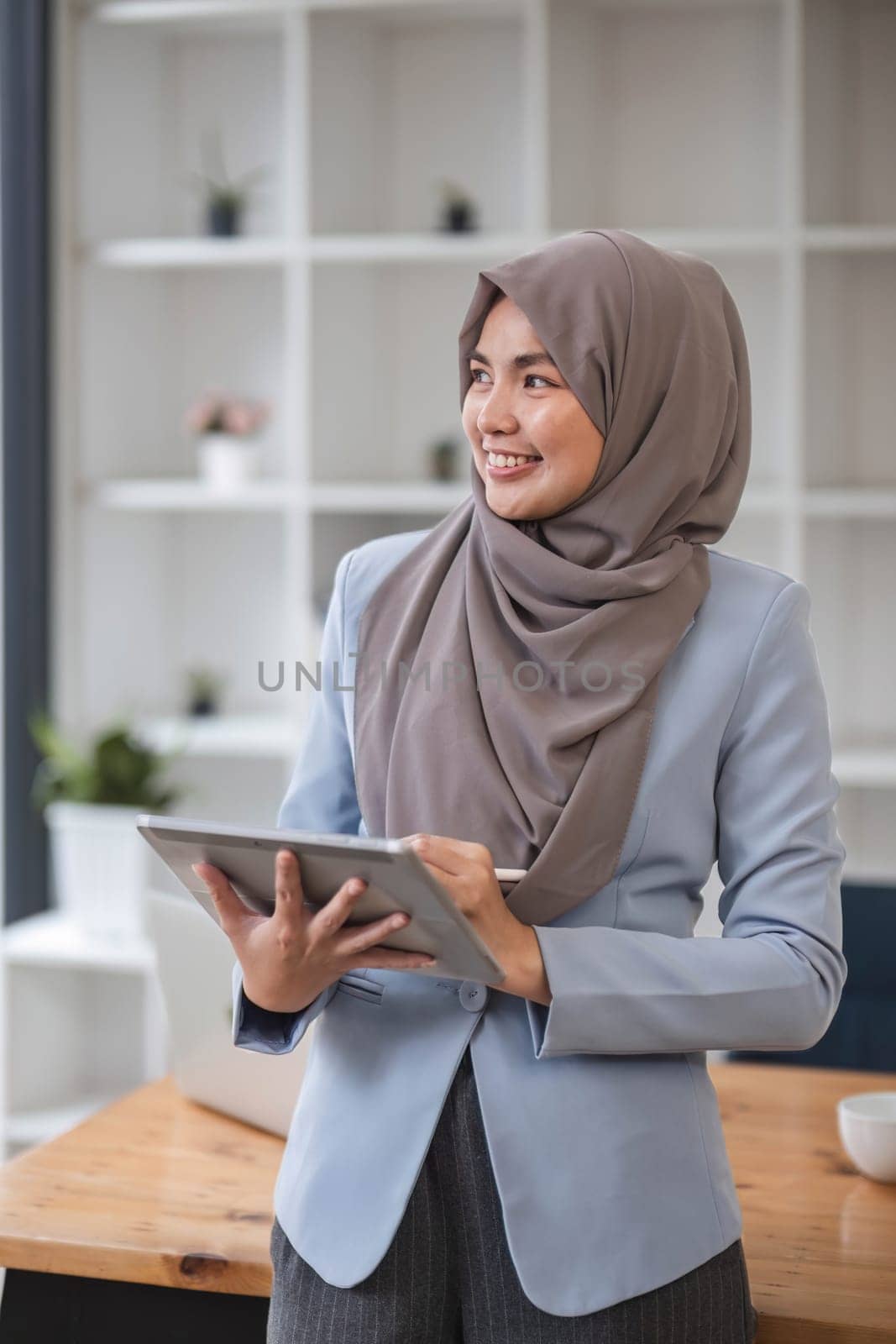 A portrait of a successful Asian Muslim businesswoman stands in her modern office with a digital tablet in her hand...