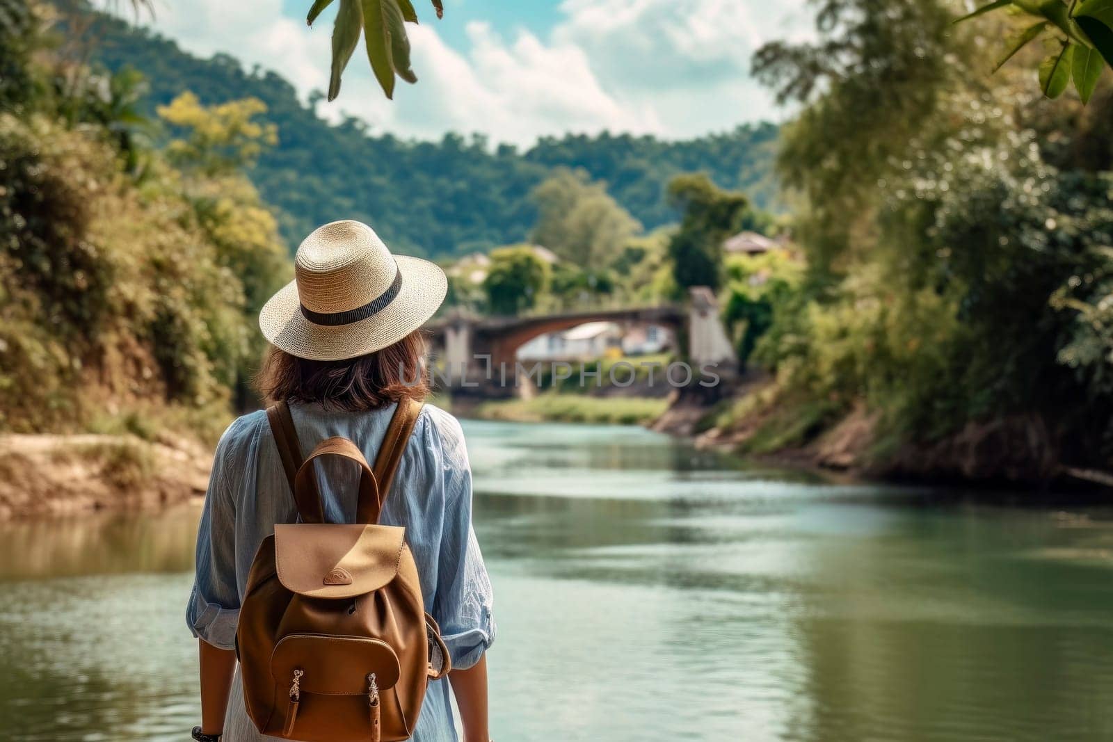 A traveling woman, with backpack and straw hat, gazes at a scenic river in a lush tropical setting.