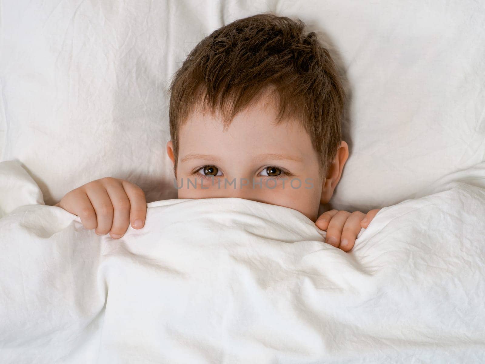 Mischief and play of young boy in bed before going to sleep. Four-year-old boy lies in bed on a pillow and looks out from under the covers