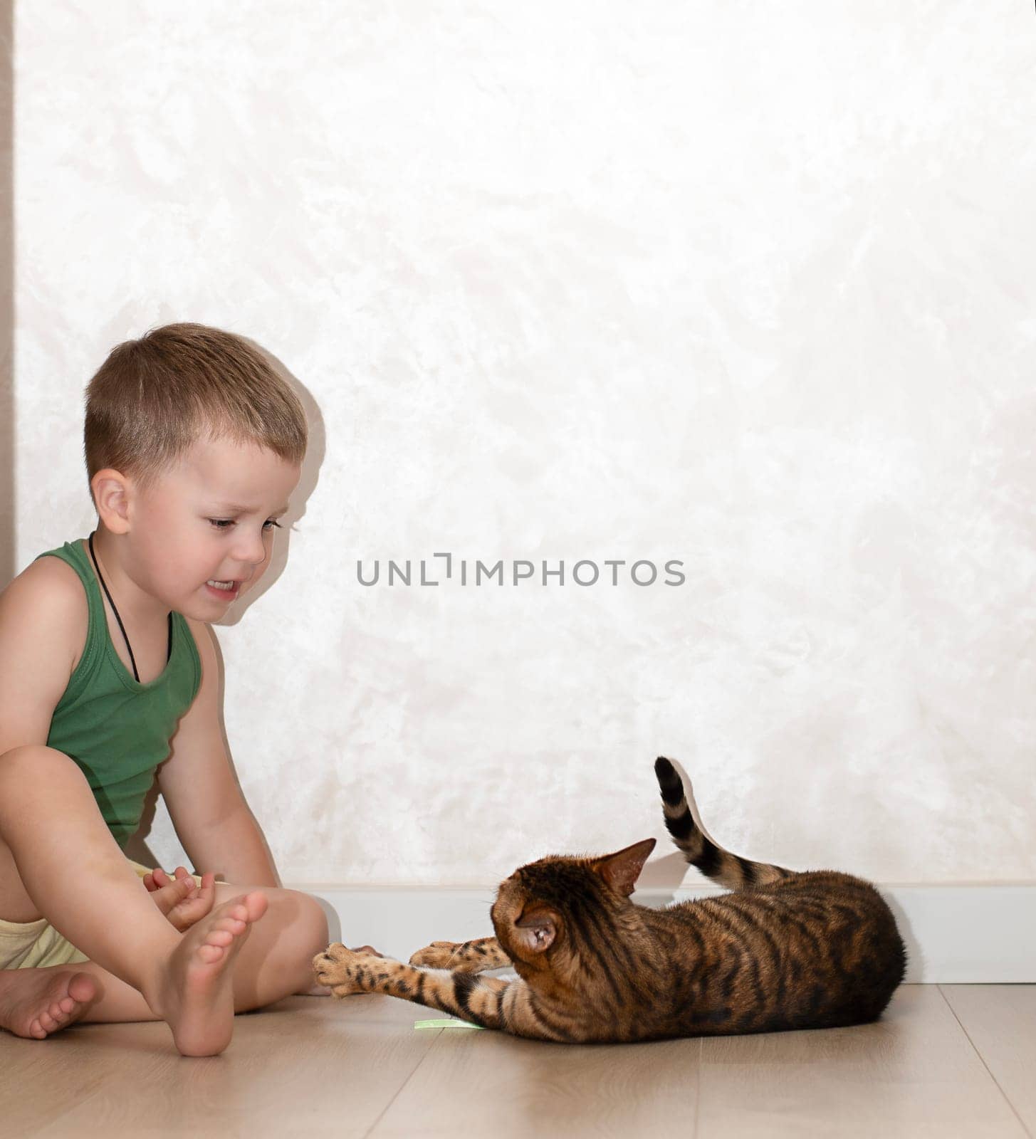 A little beautiful boy of 4 years old is having fun playing with a red, leopard bengal cat on the floor in a home interior. The domestic cat lies on the floor and plays with the baby with its paw.