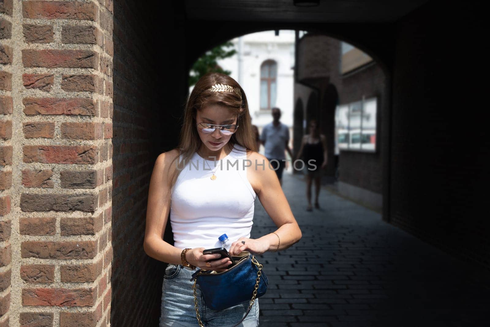 a young woman is nervously looking for something in her handbag against the background of a dark arch in a brick house,beautiful stylish clothes and gold accessories, High quality photo