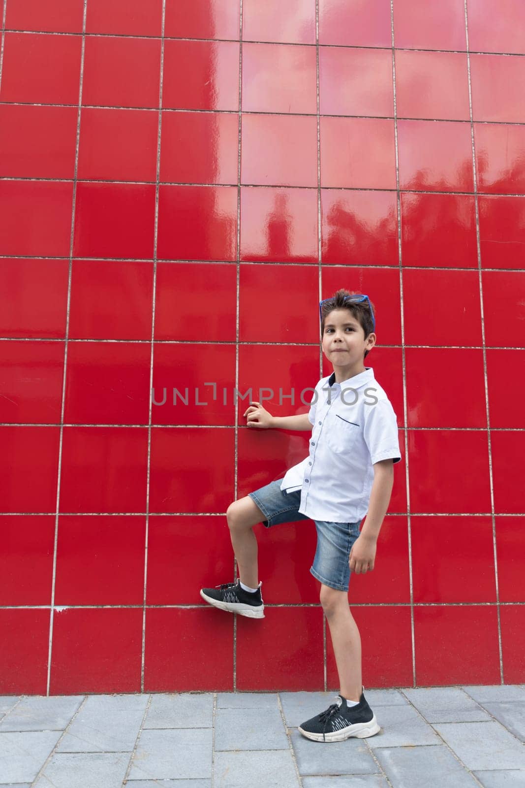 A cute boy in a white shirt raises his leg against a red wall, imitates the game of football, the child actively enjoys an active life, High quality photo