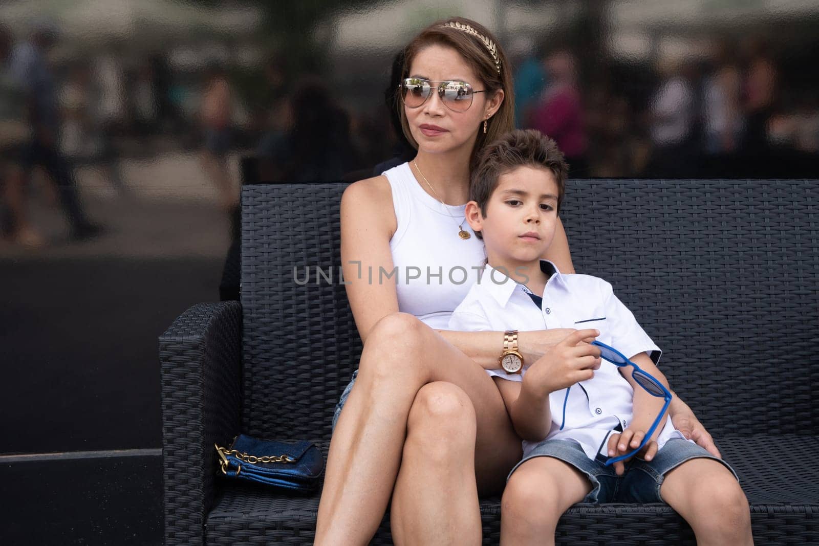 a young mother and her son in stylish clothes sit hugging on a black bench by KaterinaDalemans