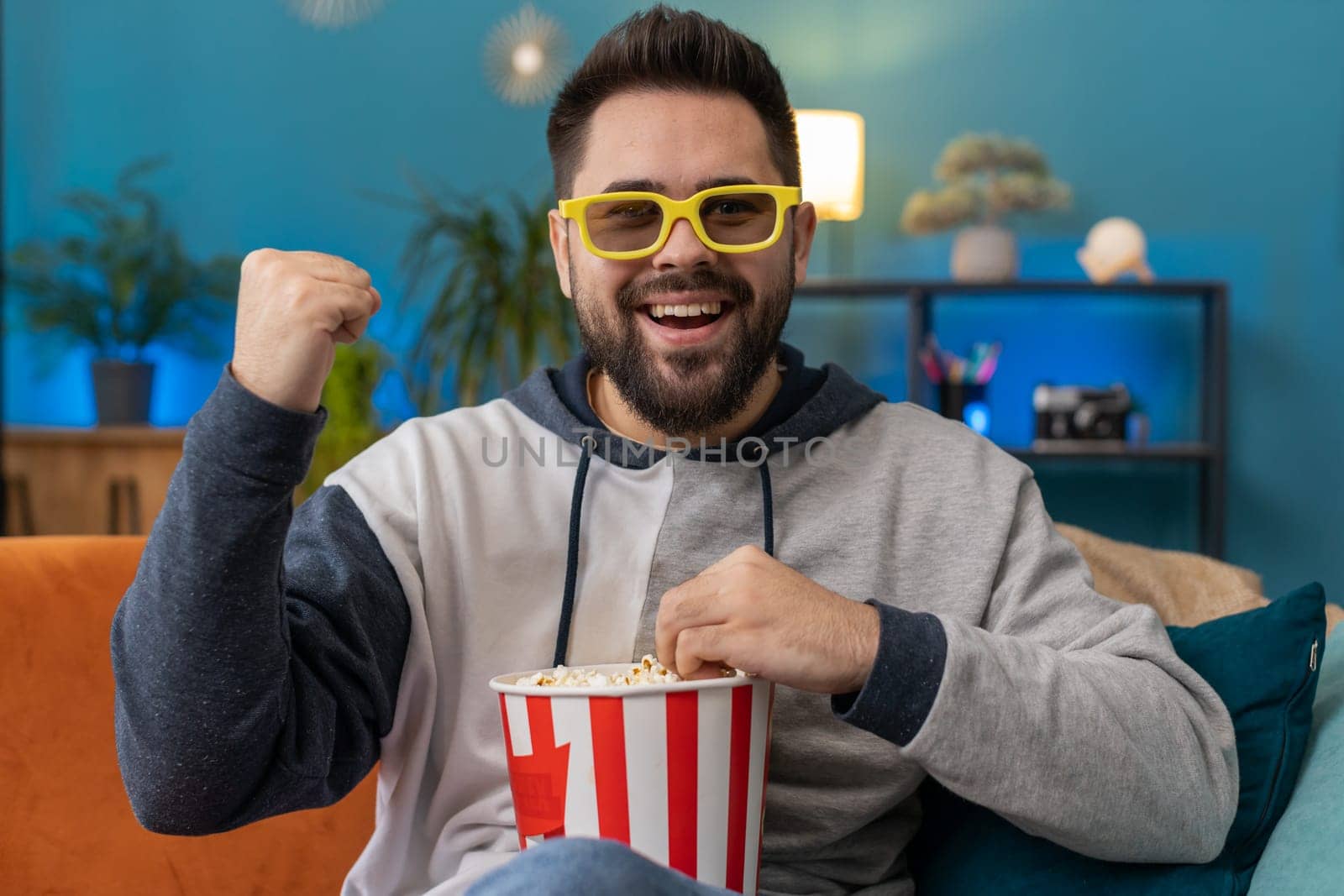 Excited young man in 3D glasses sits on sofa eating popcorn snacks and watching interesting TV serial sport game, film, online social media movie content at home apartment. Guy enjoying entertainment