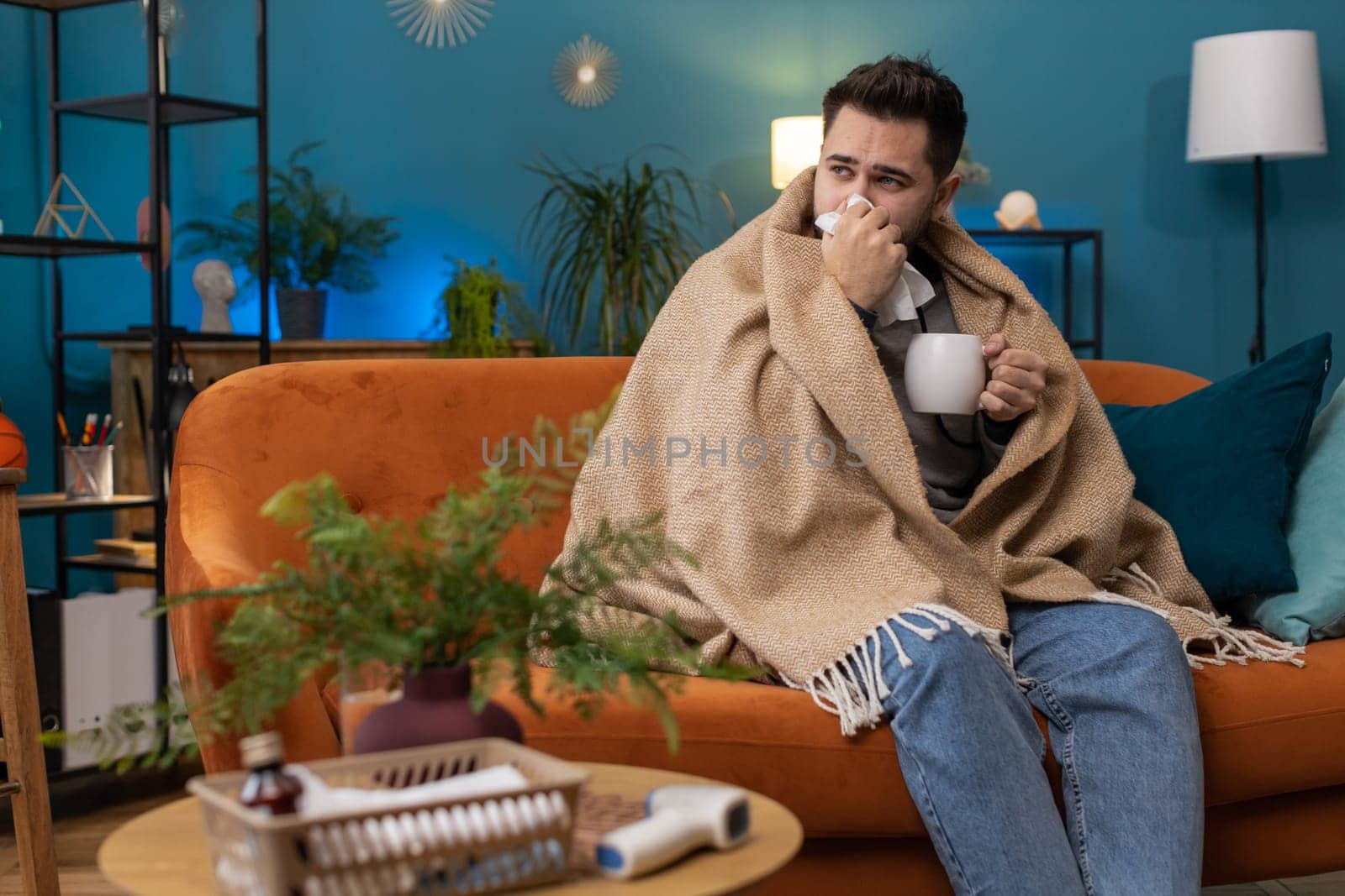 Sick man wrapped in plaid sit alone shivering from cold on sofa drinking hot tea in unheated apartment without heating due debt. Unhealthy ill guy feeling discomfort try to warming up at home room