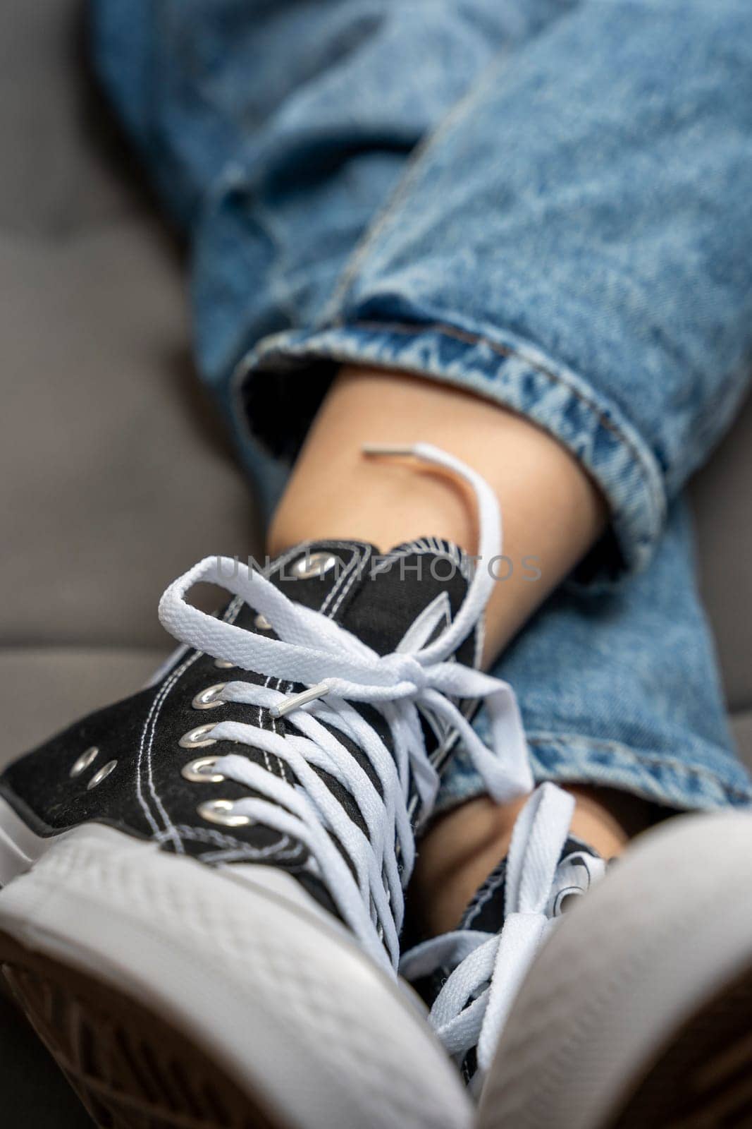 the legs of a teenage girl dressed in blue jeans and new youth sneakers with lacing lie on the home sofa. A fashionable concept in the casual style. vertical photo