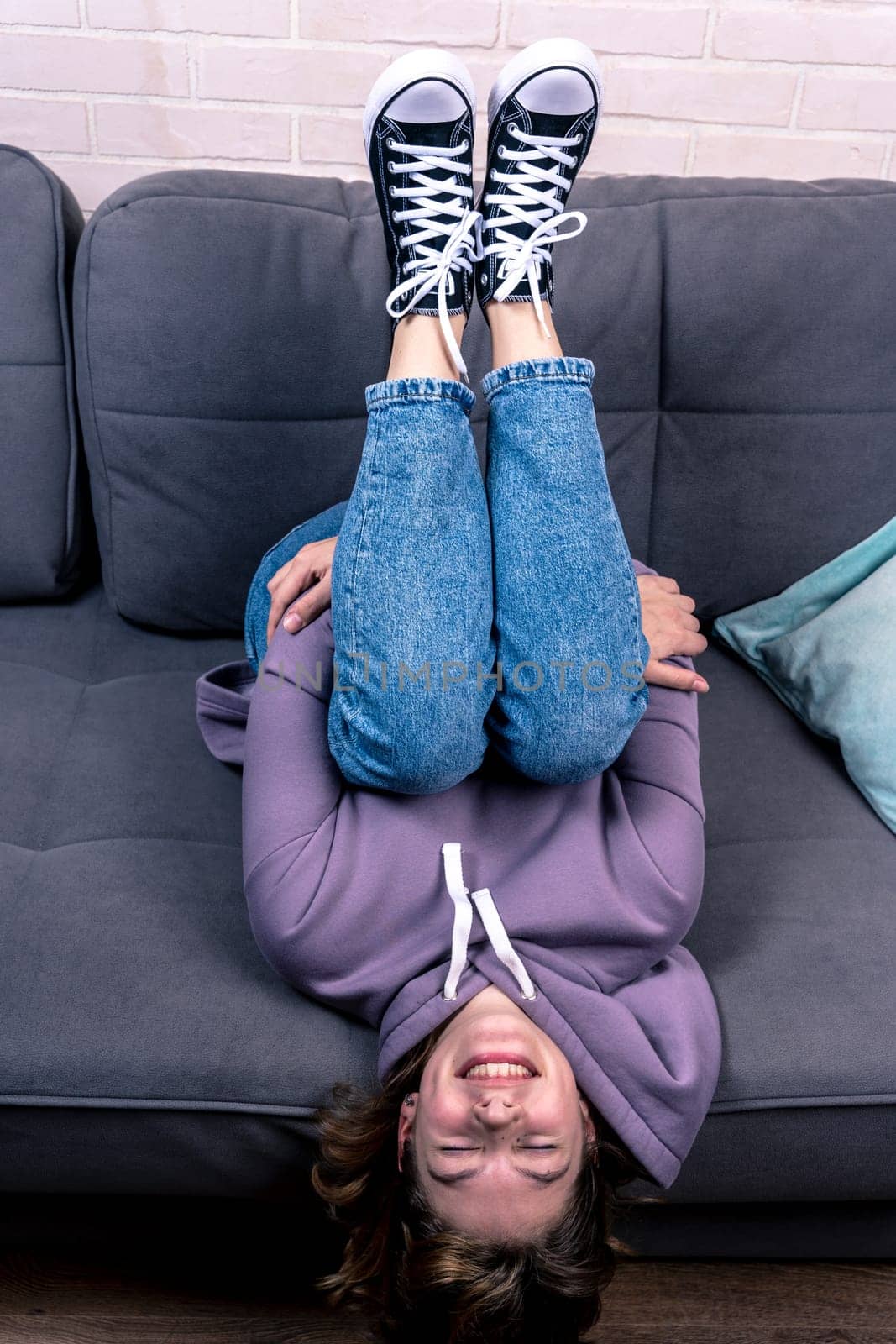 a teenage girl, lying on the couch upside down. a girl in blue jeans and hoodie sneakers relaxes at home on the couch and smiles. A fashionable concept in casual style. mental health