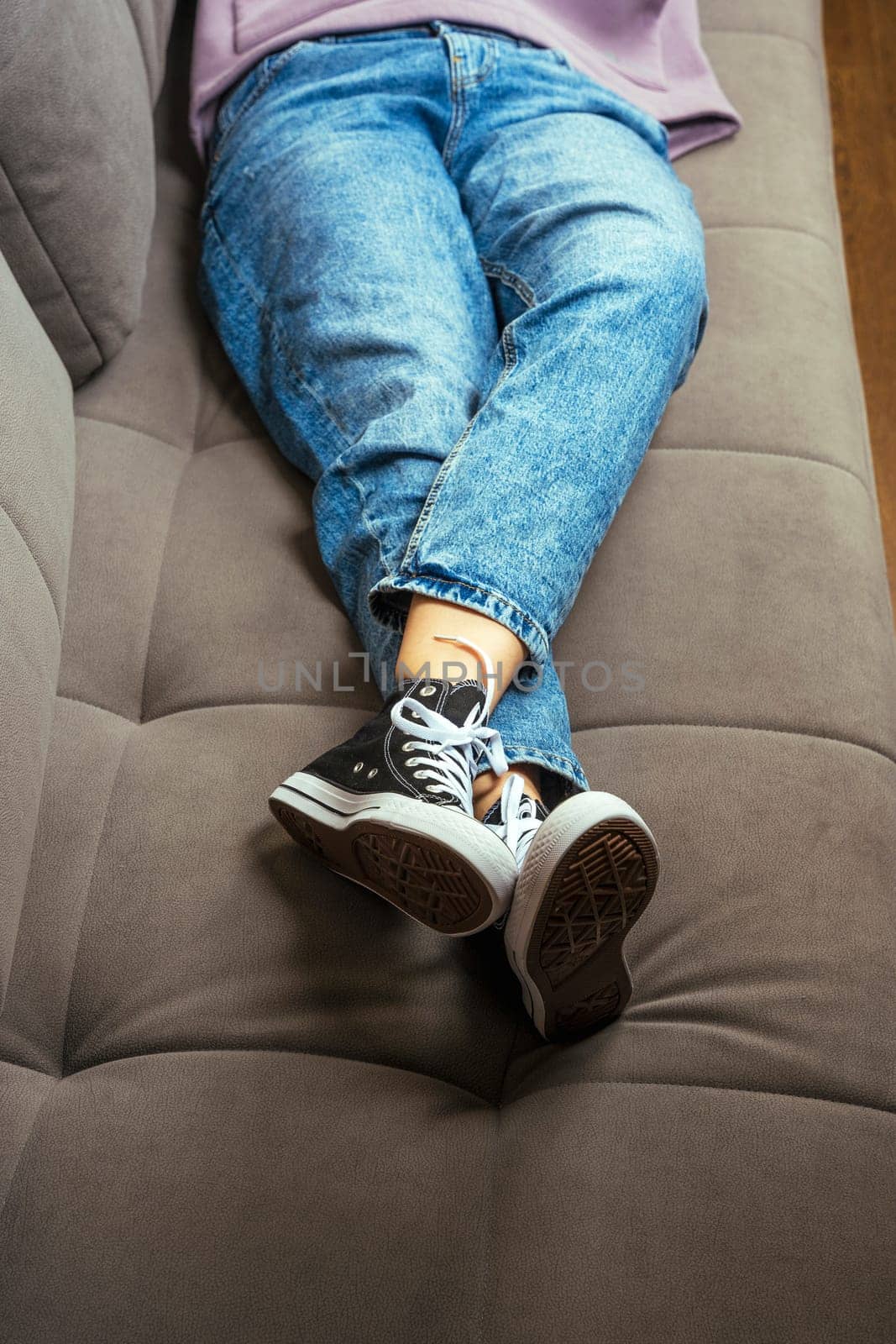 the legs of a teenage girl dressed in blue jeans and new youth sneakers with lacing lie on the home sofa. A fashionable concept in the casual style. vertical photo