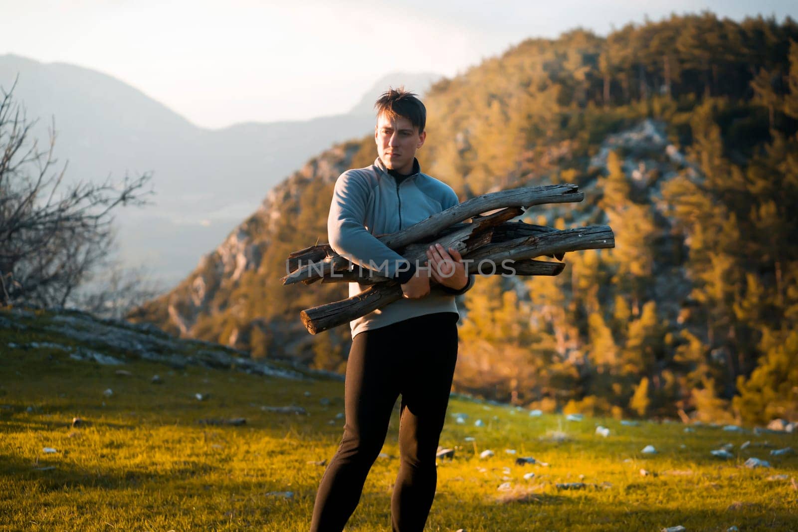 Young attractive man holds firewood, carries it to the camp against the backdrop of a picturesque landscape with hills, mountains and forest at the sunset in the evening. Traveler is hiking outdoor.