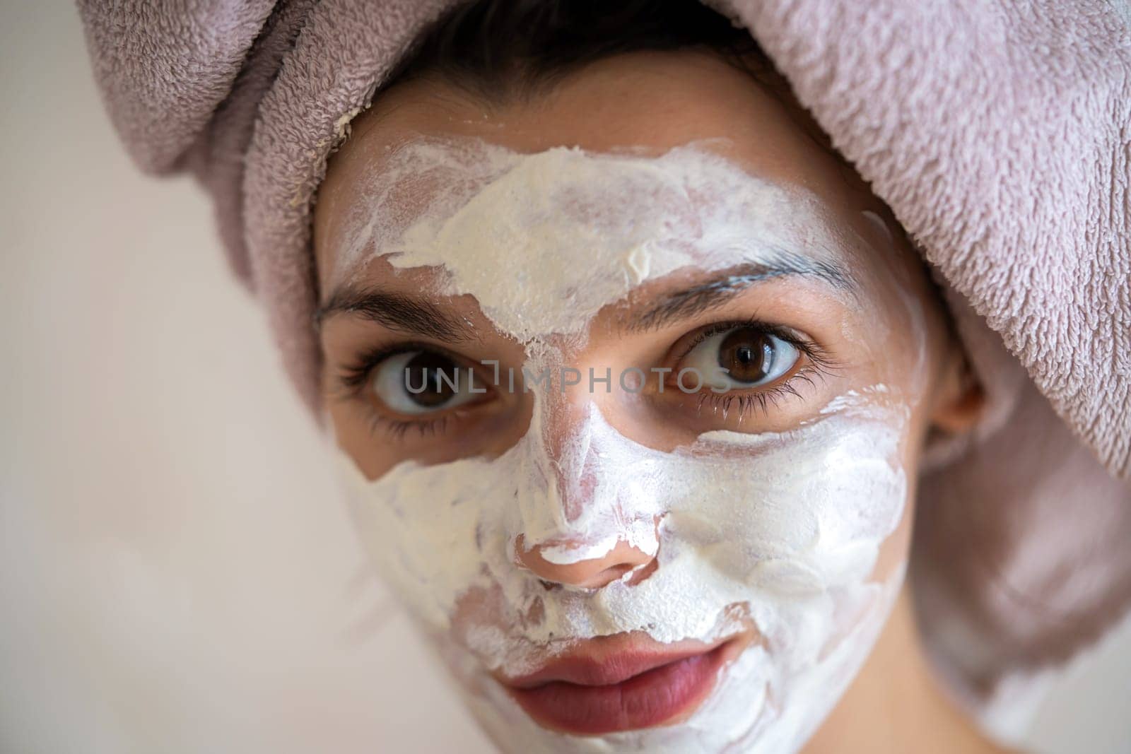 A young girl in a towel on her head and body stands near the mirror in the bathroom and applies a clay mask to her face, the woman takes care of her health and beauty.