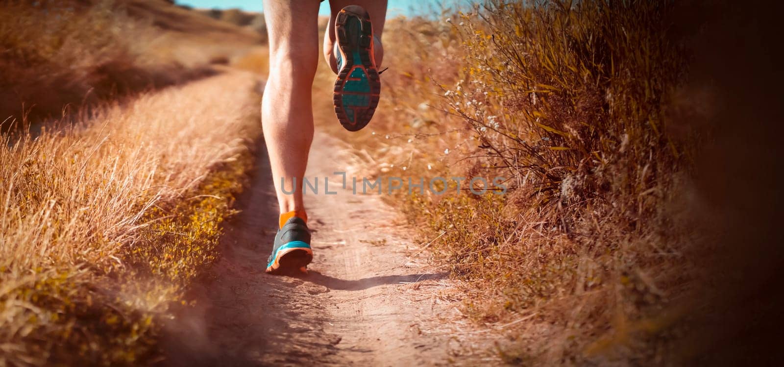 Athlete runner legs running on a close-up of a trail across a field on a shoe. Fitness and warm-up, young man with sun effect in the background.